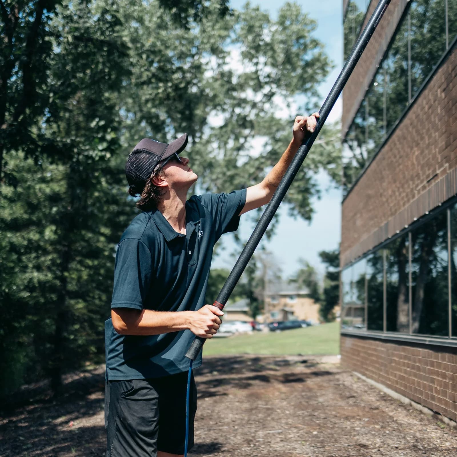 Technician reaching a water-fed pole up a brick office facade between mature trees