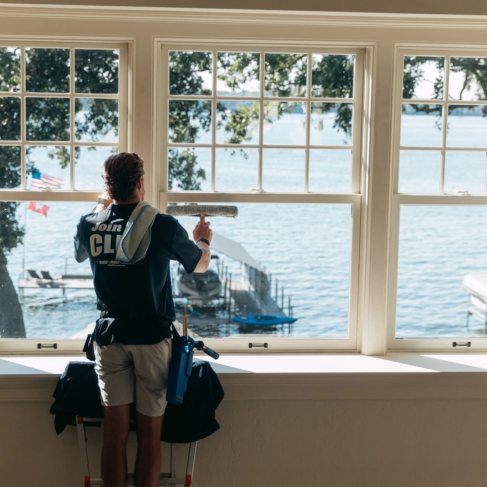 Technician cleaning a double-hung window with a lake and boats visible through the glass
